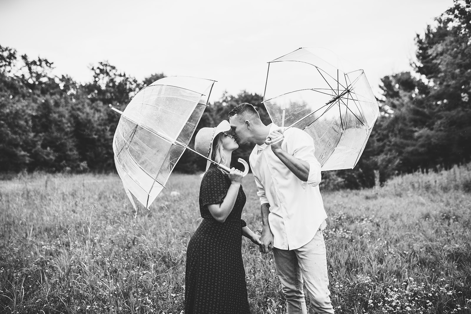 A newlywed couple shares a romantic kiss beneath two clear umbrellas, sheltered from the gentle rain. Their love radiates as they embrace, surrounded by the subtle beauty of the rainy day.