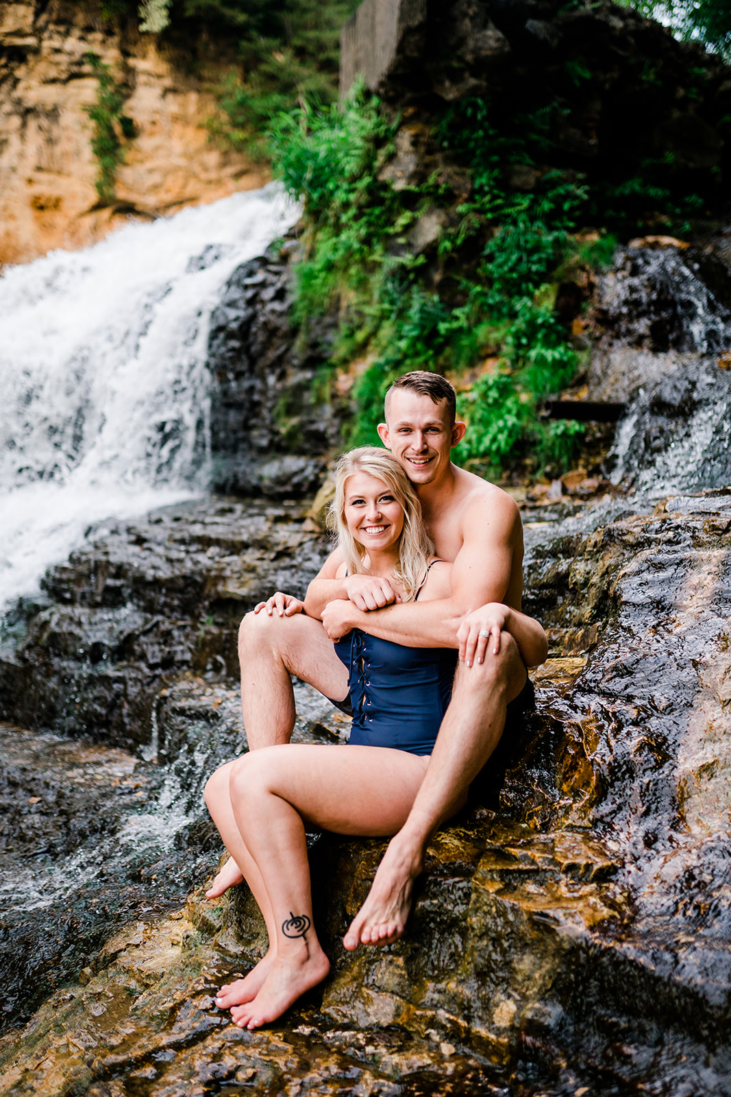 A newly engaged couple sits beside a majestic waterfall, posing for a picture together. The man tenderly wraps his arms around the woman, their smiles radiant with joy and anticipation for the journey ahead.