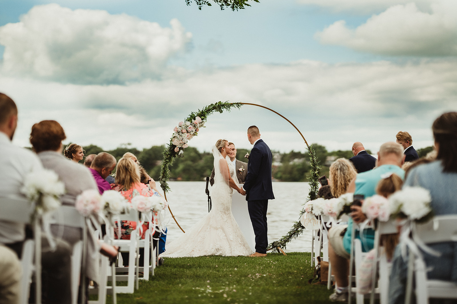 Family and friends attend a beautiful waterfront wedding ceremony on a cloudy Wisconsin day. Wedding arch inspo Ceremony photos Lakeside wedding #wisconsinweddingphotographer #choosingaphotographer #lakesidewedding #waterfrontweddingceremony