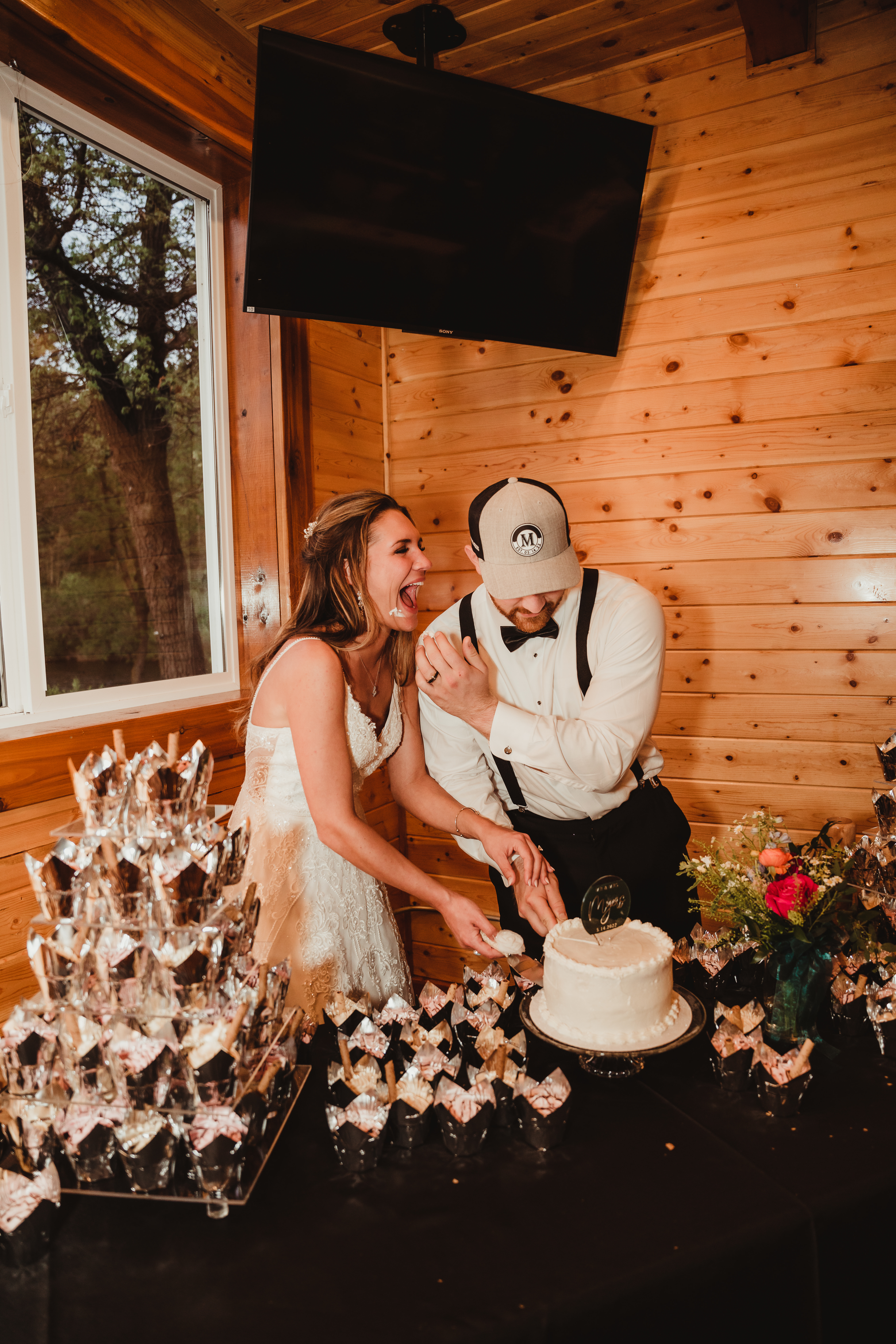 A happy bride and groom cut into their white wedding cake together at their rustic wedding reception. Wisconsin wedding reception Baseball cap groom #forestgreenwedding #rusticweddingvenue #weddingplanning #wisconsinweddingphotographer #wisconsinweddingvenue
