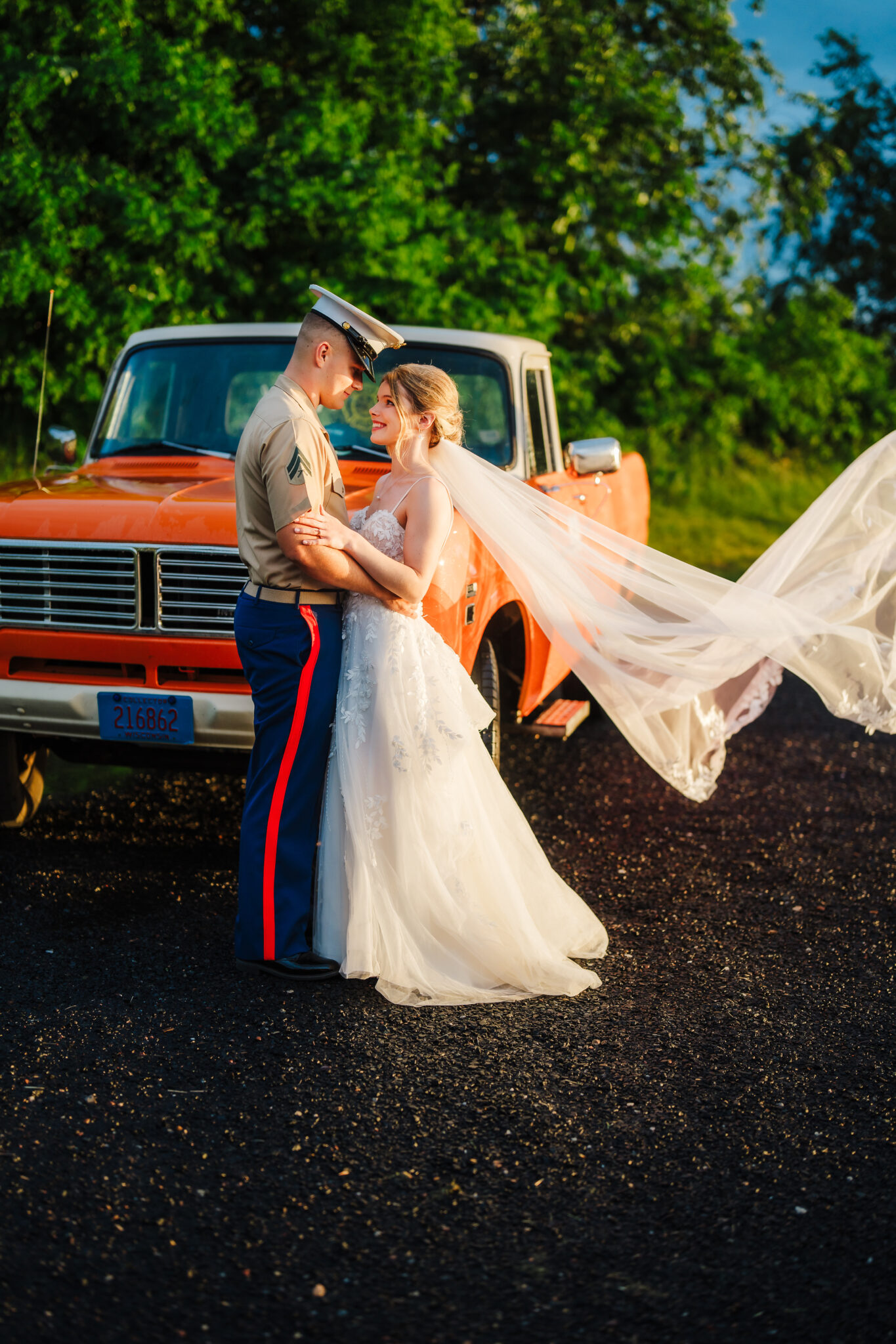 A newly married couple in a romantic embrace in front of a vintage orange truck as the bride's veil blows in the wind. 