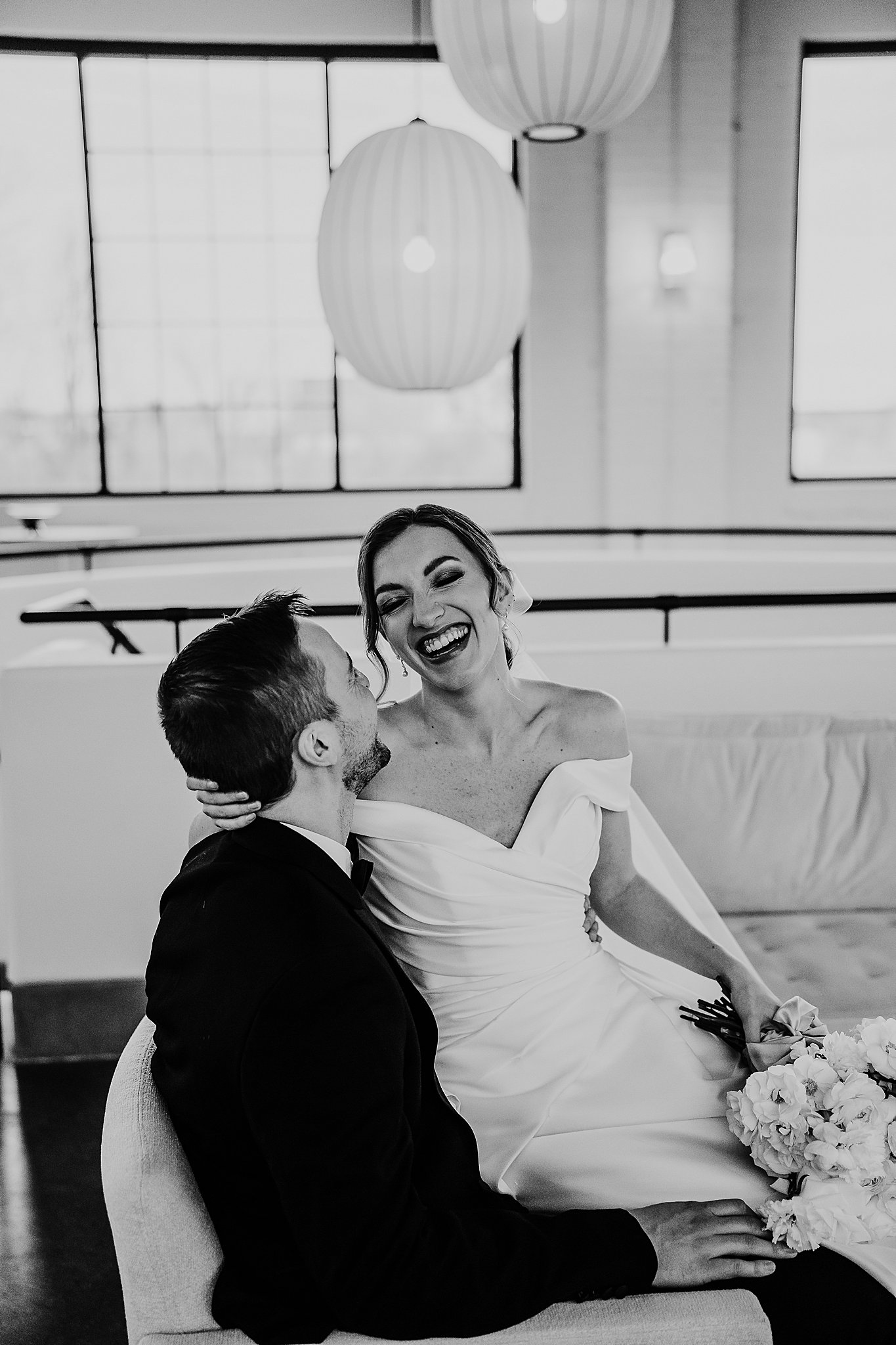 A bride laughing as her and her groom snuggle close with the bride seated on his lap. 