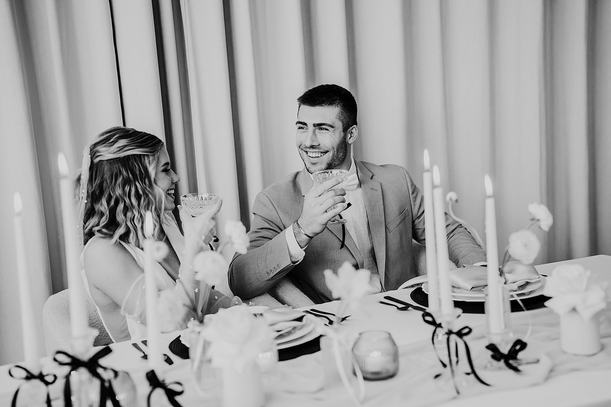 A groom takes a drink of champagne while looking at his wife and laughing. 