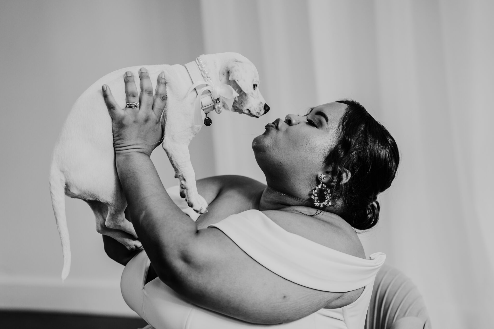 A bride raises up her puppy as she tries to kiss him. 