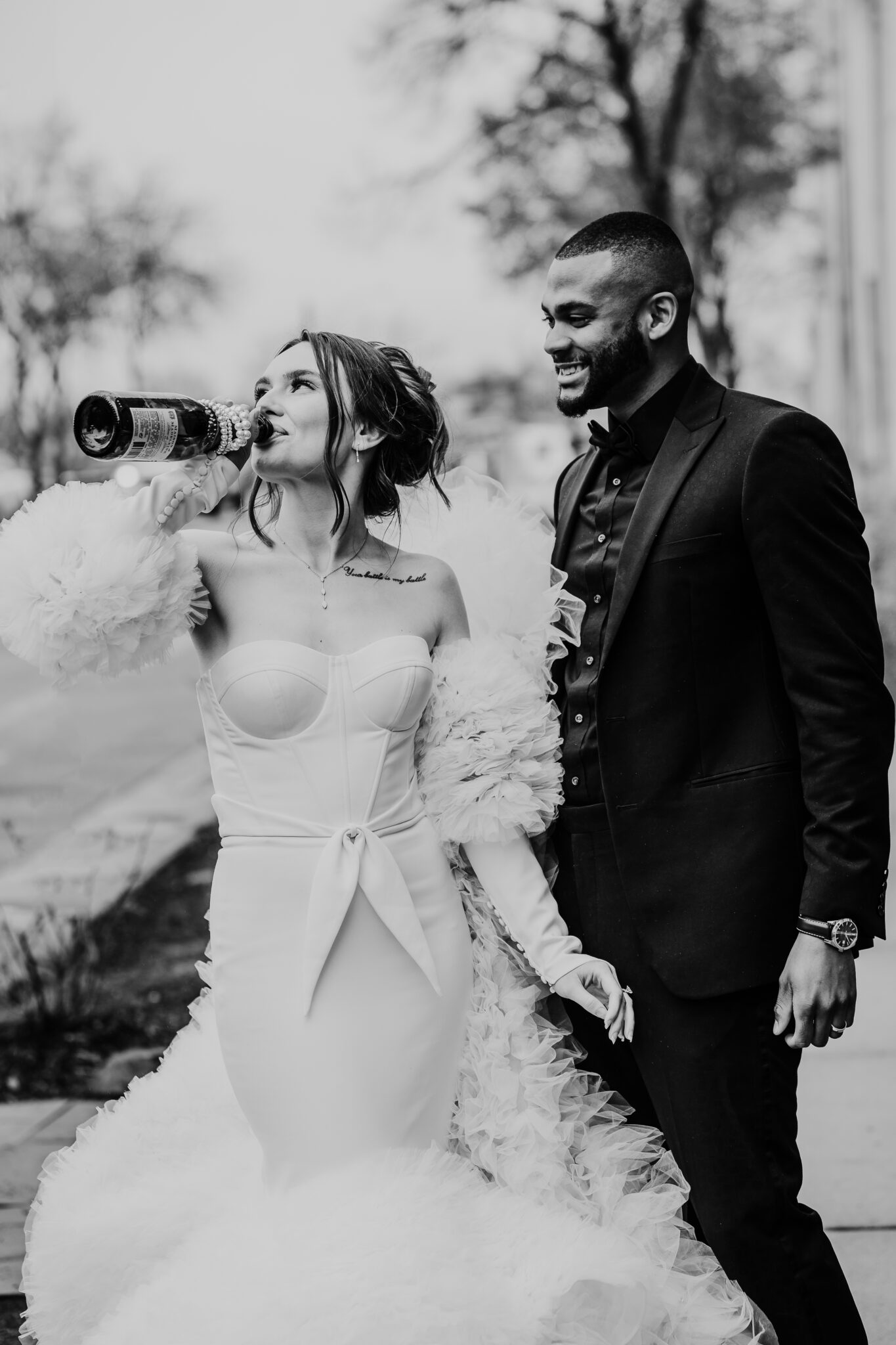 A bride takes a celebratory sip of champagne from the bottle while her groom smiles affectionately at her, capturing a moment of joy and intimacy between the newlyweds