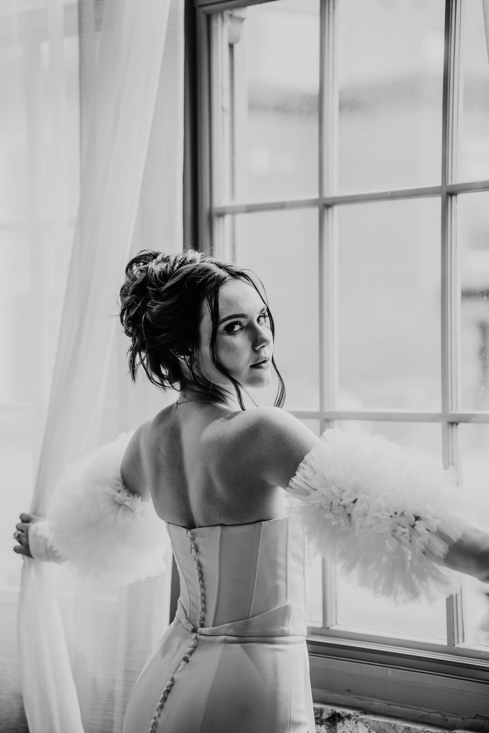 A stunning bride stands in front of a window, looking directly at the camera with a serene expression, exuding elegance and confidence. Her poised demeanor and captivating gaze create a timeless and captivating portrait.