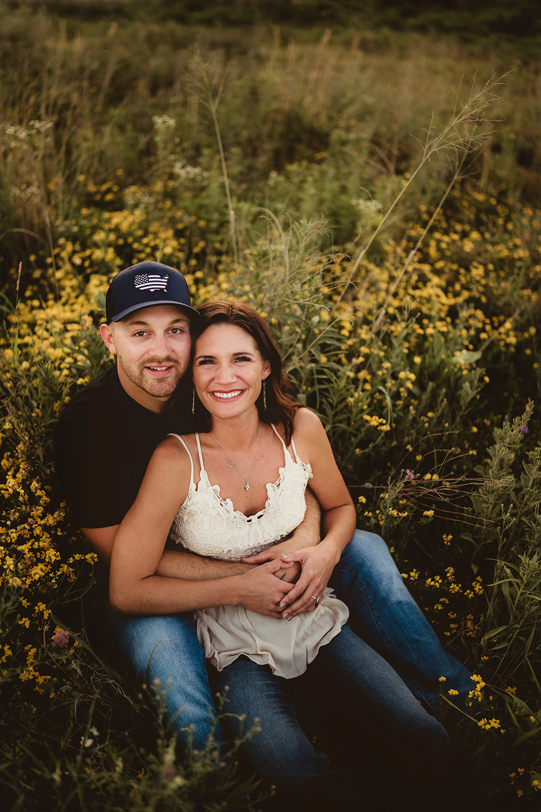 A happy engaged couple snuggles close, smiling at the camera amidst a beautiful flower field.