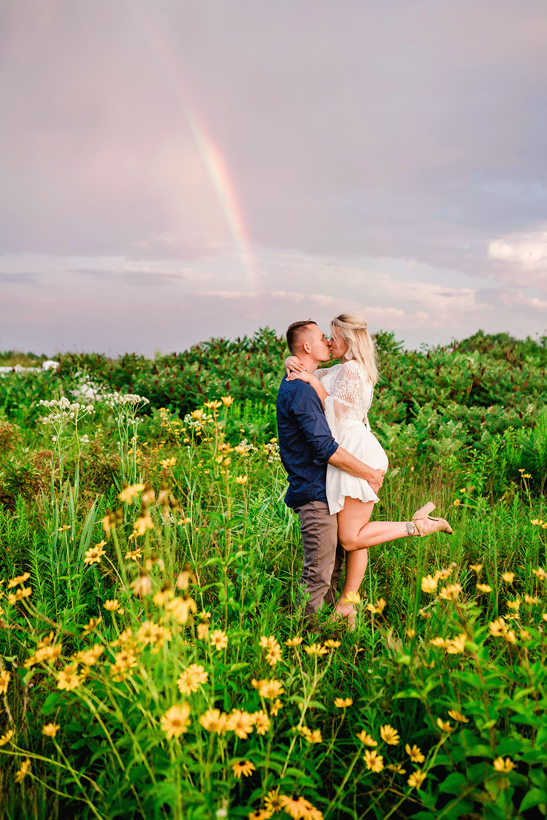 n a field of vibrant yellow flowers under a radiant rainbow, a man lifts his partner tenderly, sharing a loving kiss, capturing a moment of pure joy and romance.
