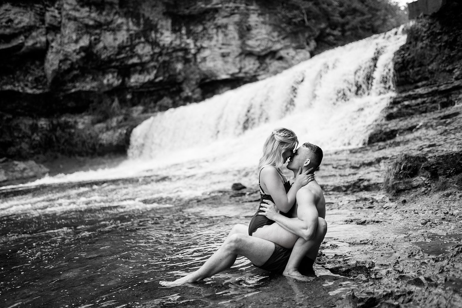 A couple shares a romantic kiss while laying in the river, framed by a majestic waterfall in the background. The serene beauty of nature enhances the intimacy of the moment