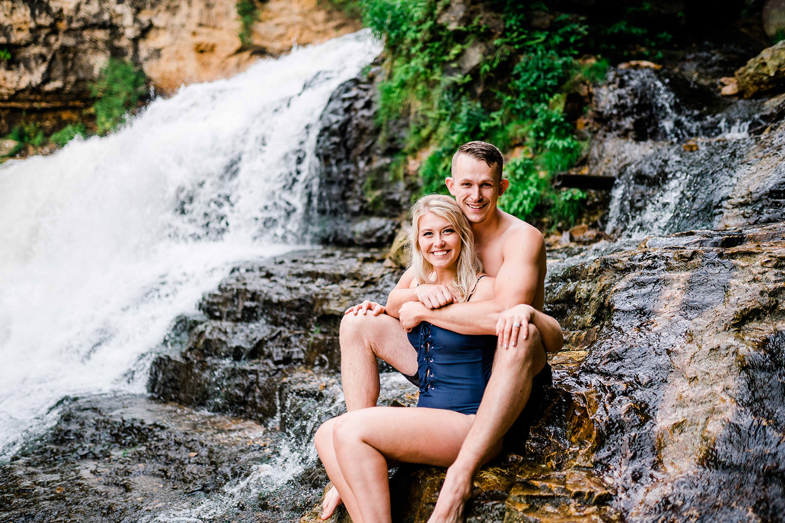 A smiling couple sits beside a picturesque waterfall in Wisconsin, their happiness evident as they pose for the camera against the backdrop of nature's beauty.