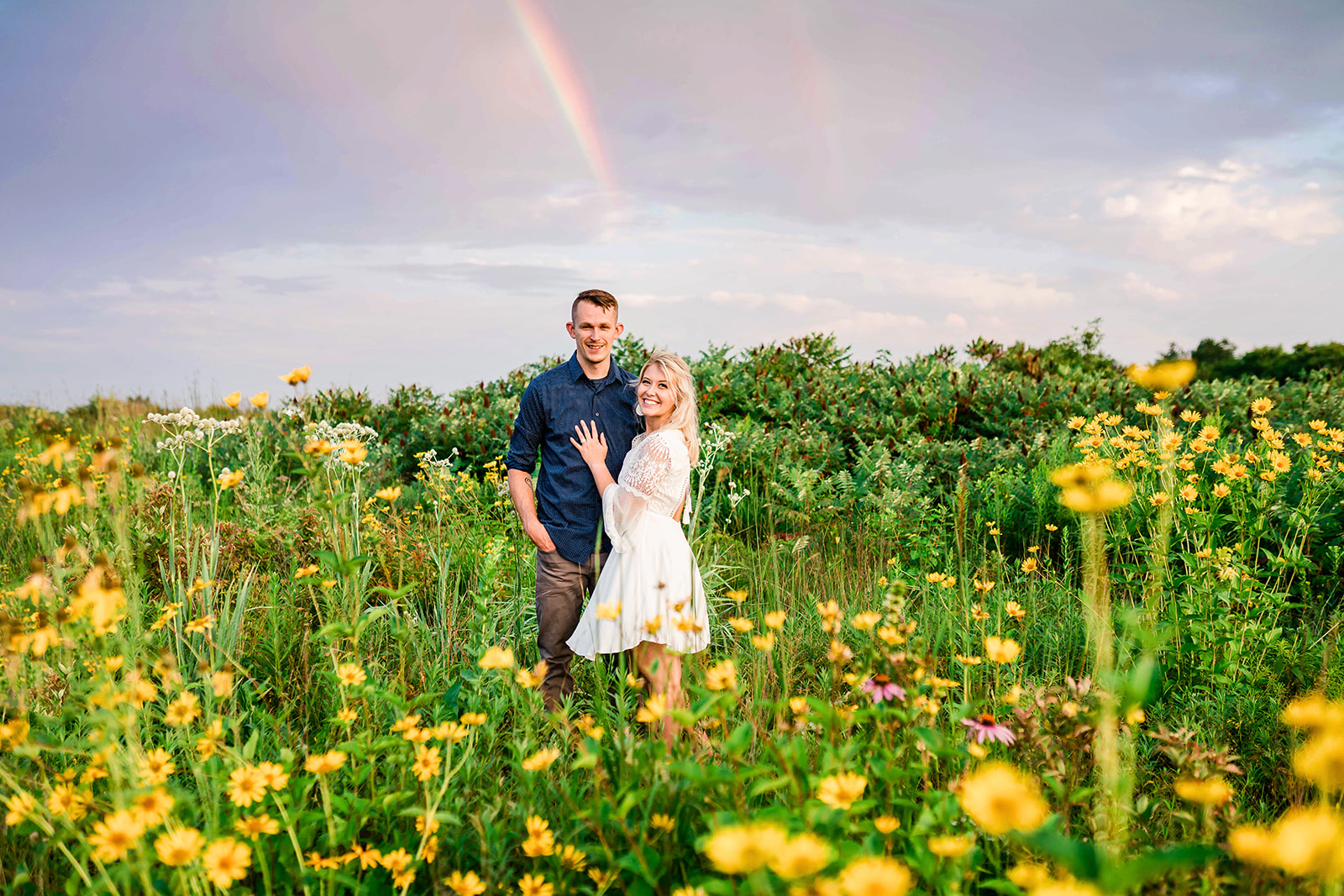 A joyful engaged couple smiles at the camera under a vibrant rainbow in a lush flower field.