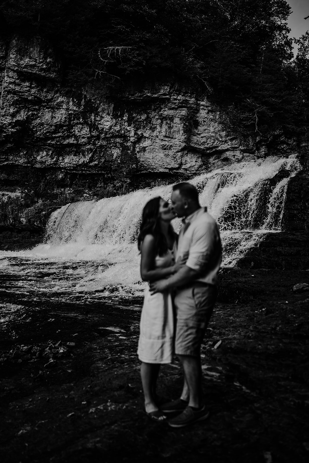 An affectionate engaged couple shares a kiss with a majestic waterfall in focus behind them.