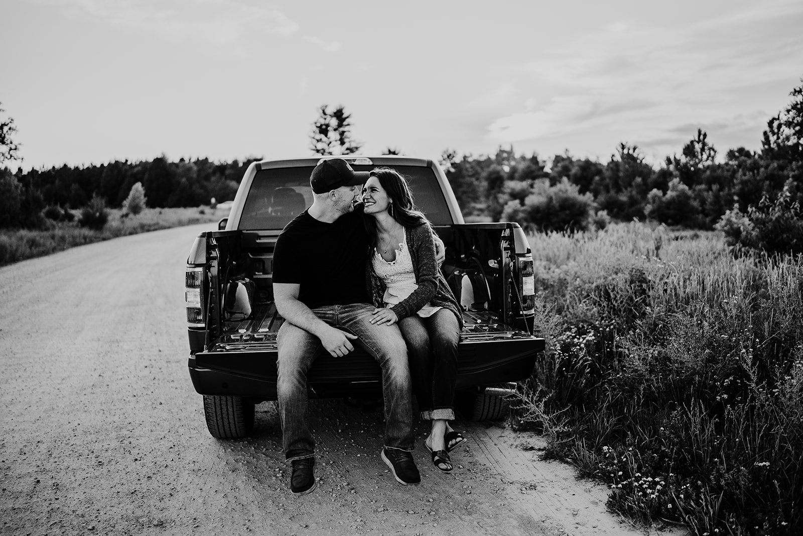 A happy couple sits in the bed of their truck, sharing smiles and affectionate glances with each other, creating a moment of intimacy amidst their outdoor adventure.