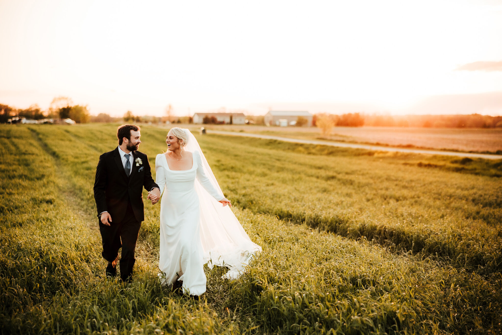 A couple runs joyfully through a field with a rustic barn in the background, silhouetted against a vibrant sunset.
