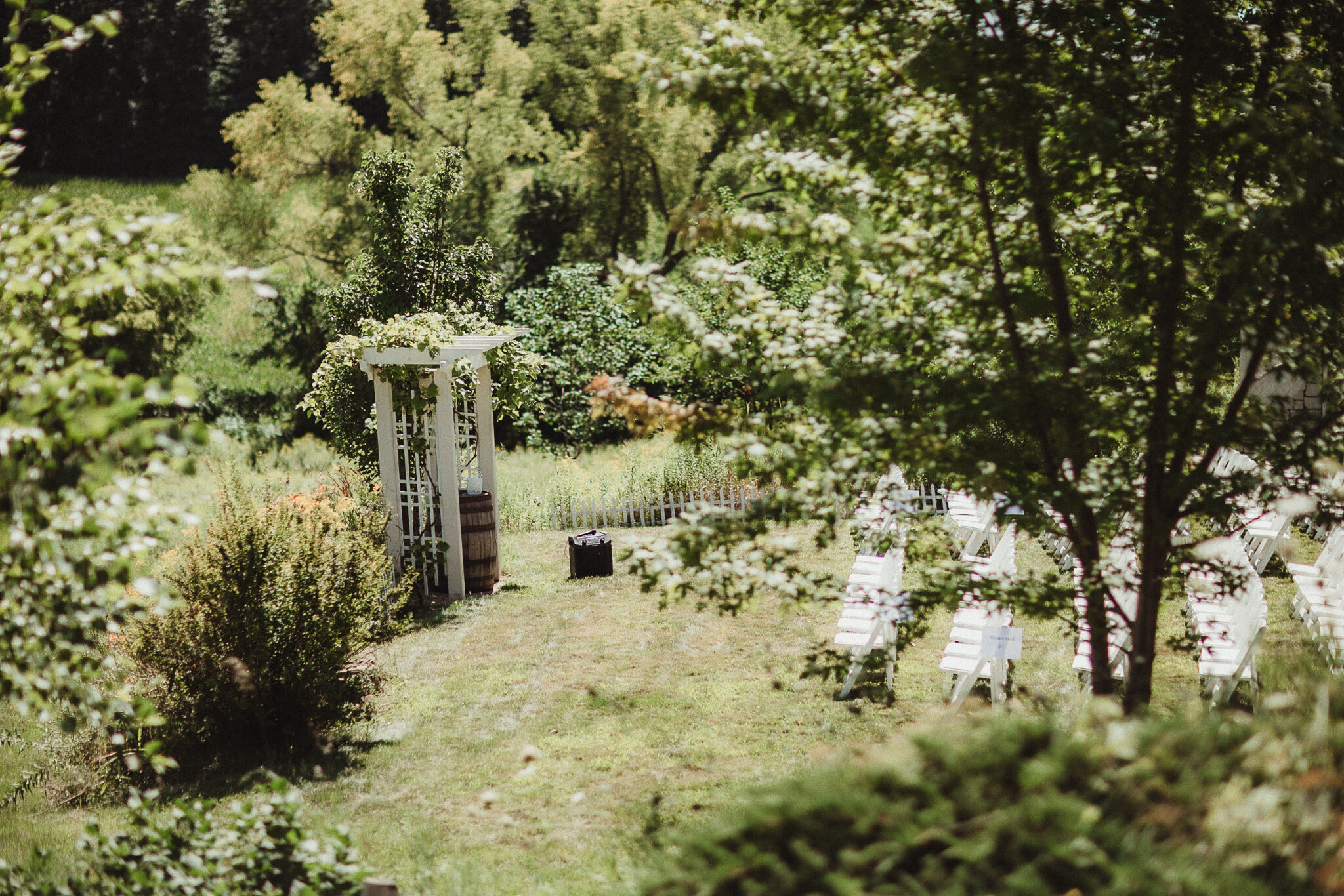 A sunlit outdoor wedding ceremony space with rows of white chairs adorned with delicate floral arrangements, facing an arch draped in greenery and blooms.