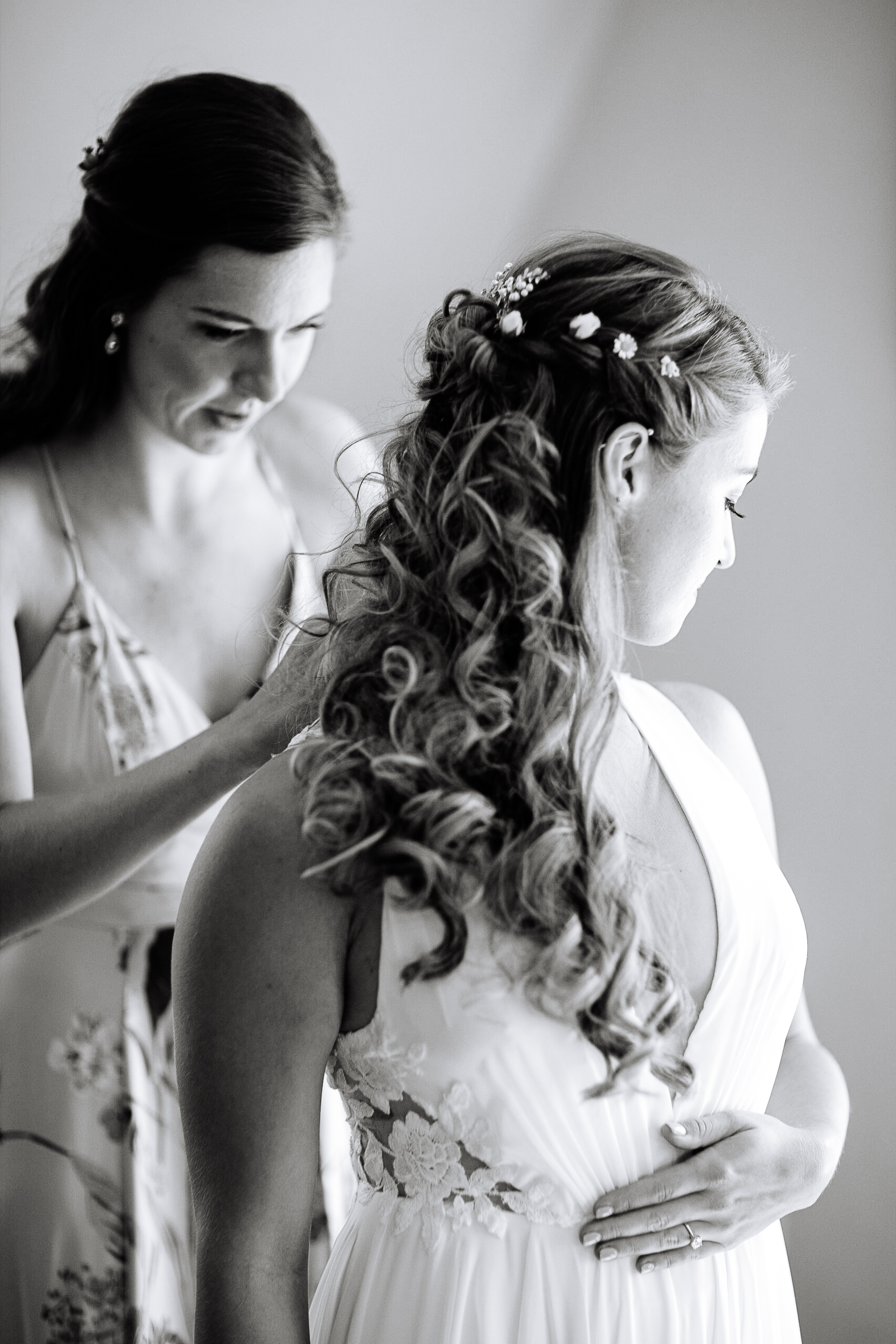 A black-and-white image of a bride with intricate curls adorned with delicate floral pins as a bridesmaid adjusts her dress. The bride’s lace wedding gown features elegant floral embroidery, and her hand rests gently on her waist, showcasing her engagement ring. The moment captures quiet preparation and heartfelt connection before the ceremony." Let me know if you'd like to refine it further